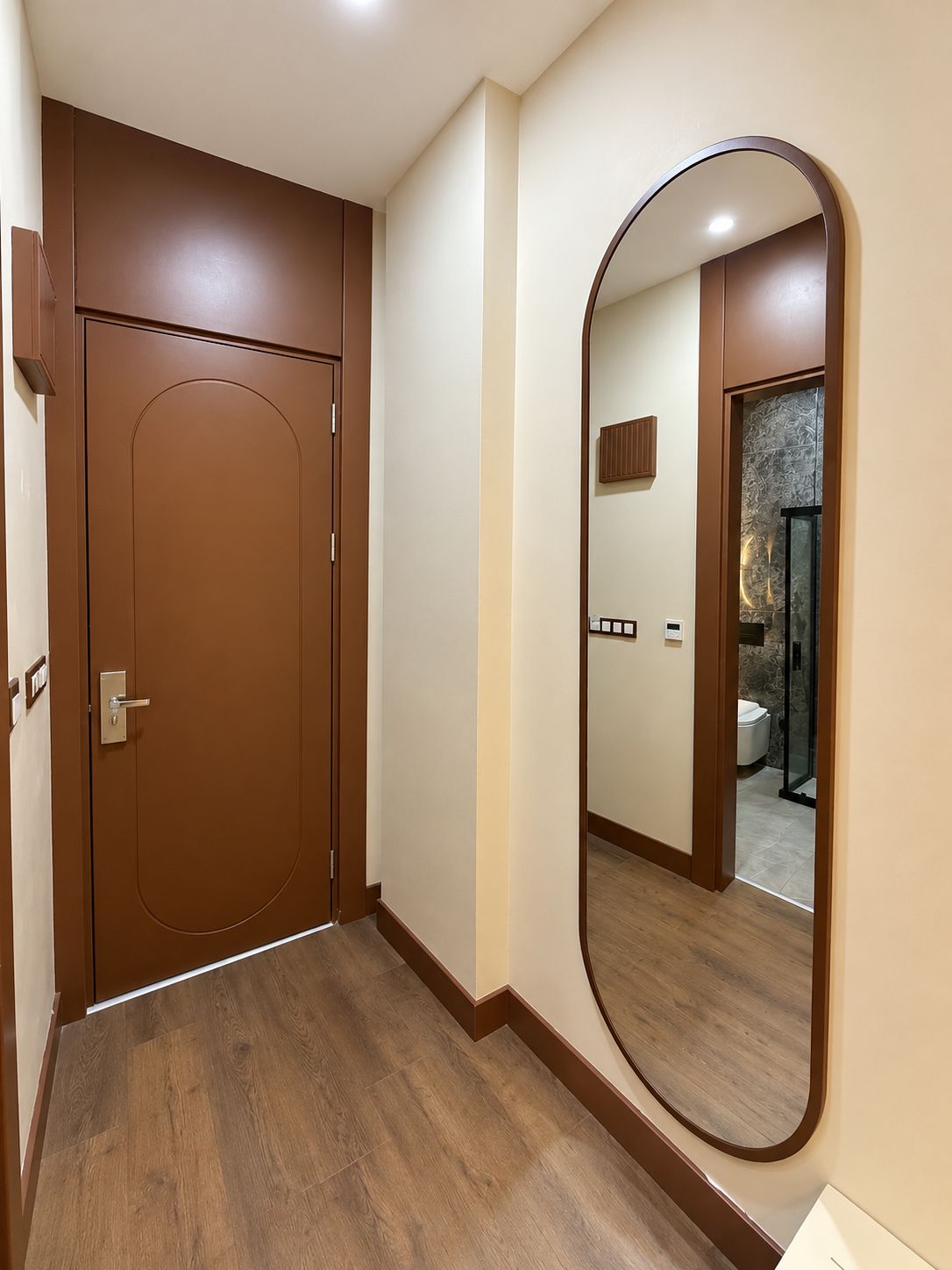 A hallway with a wooden door and a large mirror reflecting a bathroom with a shower and toilet.
