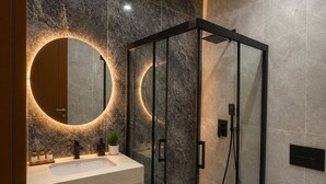 A modern bathroom with a glass-enclosed shower, a round mirror with a light, a white sink, and a toilet.