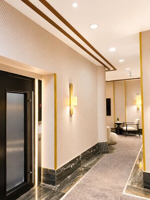 Un pasillo moderno de hotel con suelo de mármol, luces empotradas en el techo y una puerta de ascensor negra.