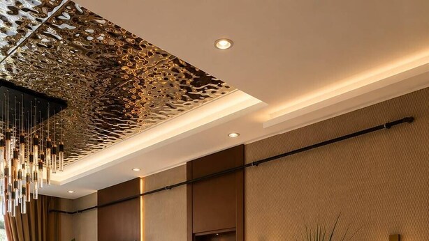 A hotel lobby with a modern design, featuring a patterned ceiling, a flat-screen TV, seating area with chairs and tables, and a large potted plant.