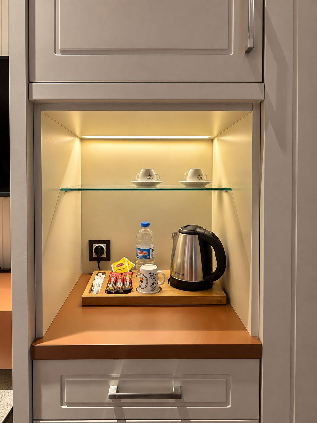 Eine Minibar mit einer Glasablage, zwei Tassen, einer Wasserflasche, einer Kaffeemaschine und kleinen Snacks.