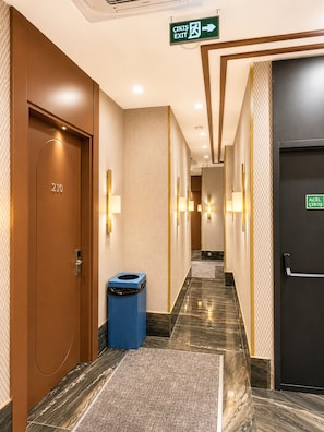 A hotel corridor with a green exit sign, a blue trash bin, and a door labeled '210'. - Ahura Otel (Diyarbakir)