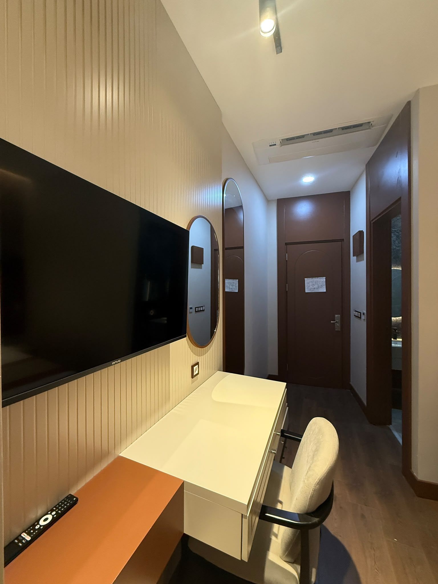 Ein Hotelzimmer mit einem Flachbildfernseher, einem Nachttisch mit Fernbedienung, einem Sessel, einem Spiegel und einer Tür zu einem anderen Raum.