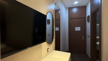 Ein Hotelzimmer mit einem Flachbildfernseher, einem Nachttisch mit Fernbedienung, einem Sessel, einem Spiegel und einer Tür zu einem anderen Raum.