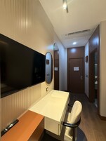 Habitación de hotel con televisor de pantalla plana, mesita de noche con control remoto, una silla, un espejo y una puerta que da a otra habitación.