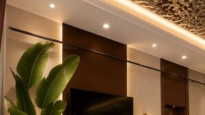 A modern hotel lobby with a large potted plant, two armchairs, a small table, and a flat-screen TV mounted on the wall.