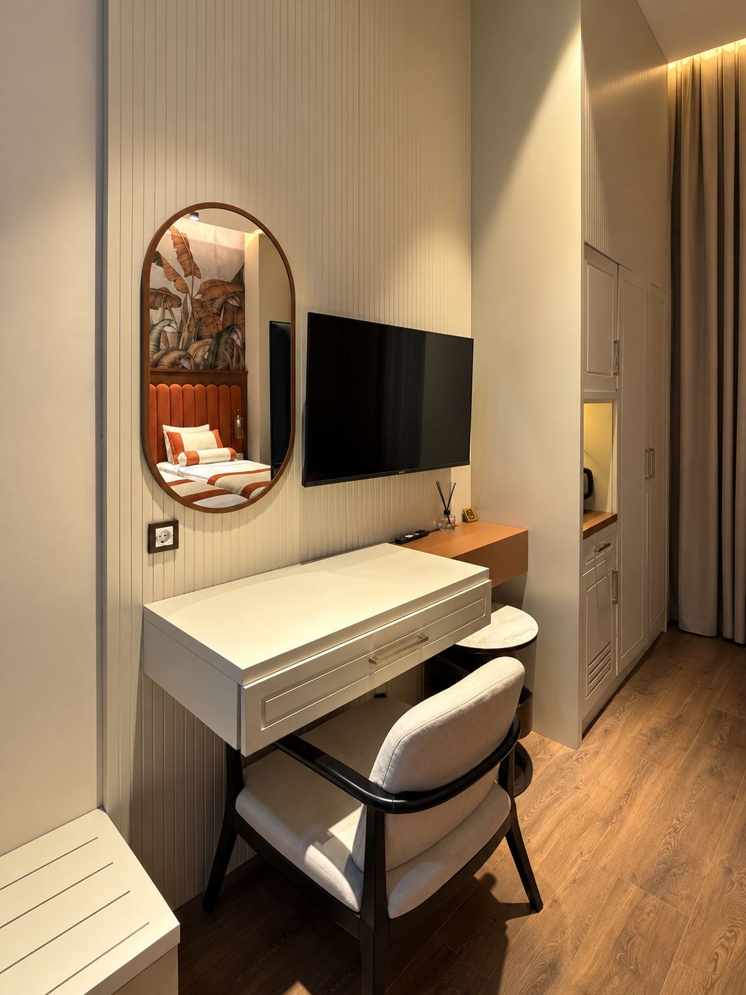 A modern hotel room with a white desk, a flat-screen TV, a mirror, a bed with a red headboard, and a wooden floor.