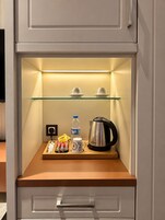 Un minibar con repisa de vidrio, dos tazas, una botella de agua, una tetera y algunos bocadillos.