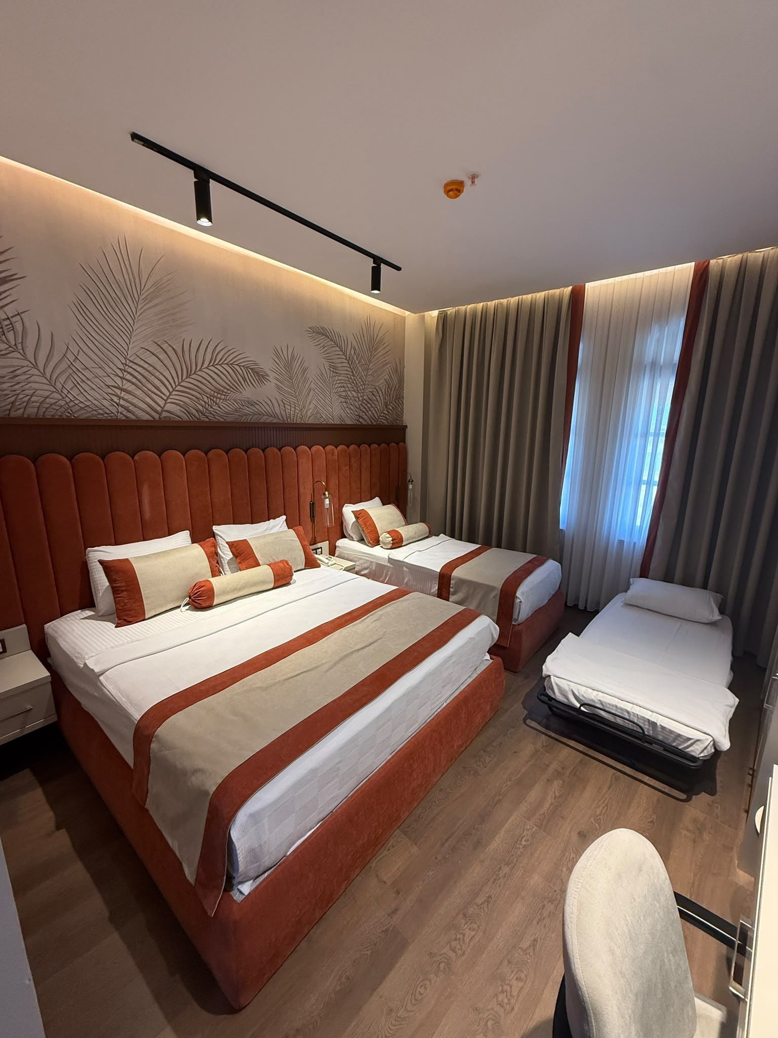 Ein Hotelzimmer mit zwei Betten, Holzboden, einem großen Fenster mit Vorhängen und einer Wand mit einem Blattmuster.