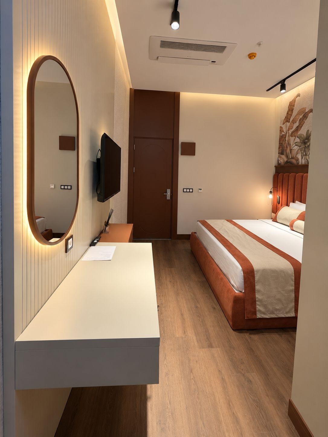 A modern hotel room with a large bed, a bedside table, a flat-screen TV, a mirror, and a ceiling-mounted air conditioning unit.