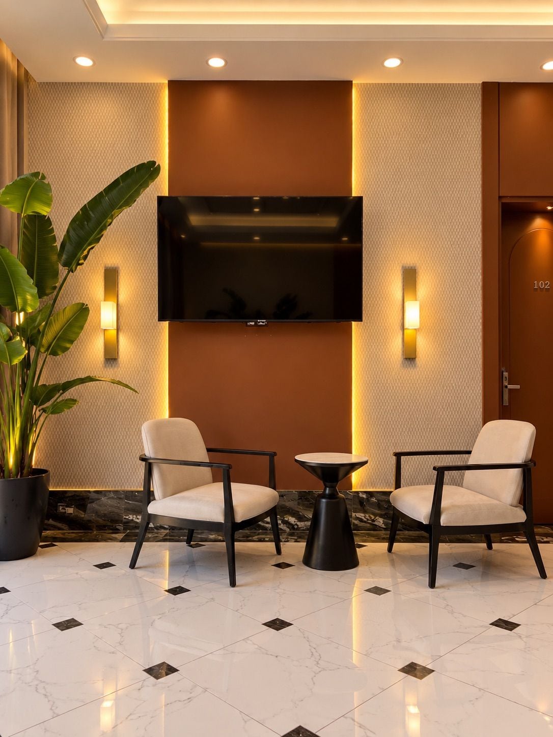 Um hall de entrada com duas poltronas, uma mesa pequena, um vaso de planta, uma TV de tela plana na parede e uma porta com o número 102.