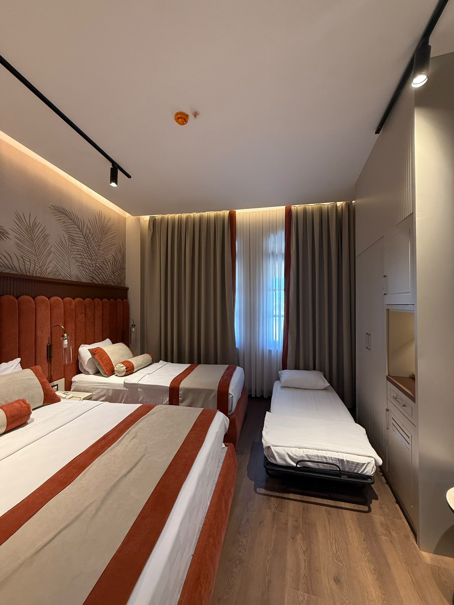 Ein Hotelzimmer mit zwei Betten, Holzboden, großem Fenster mit Vorhängen und einem am Wand befestigten Fernseher.