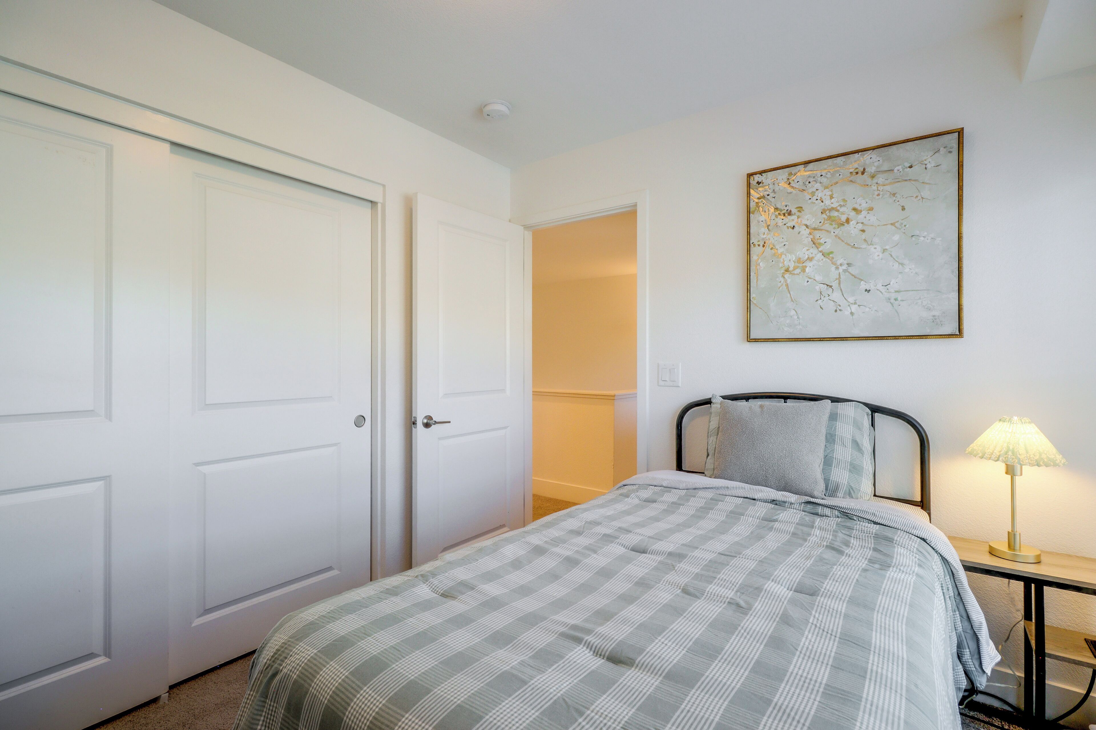 A bedroom with a bed, a nightstand with a lamp, a door, and a wall-mounted painting.