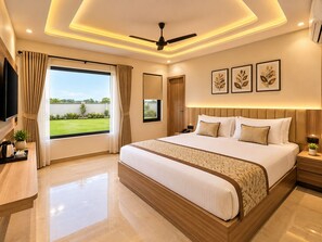 A modern bedroom with a large bed, a ceiling fan, a TV, and a view of a green landscape. - Hotel Akrati Inn Vrindavan (Mathura)