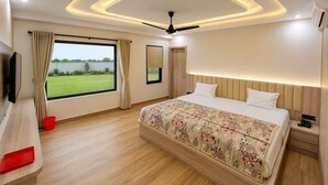 Un dormitorio con cama, televisión, ventana con cortinas, ventilador de techo y vista a un paisaje verde.