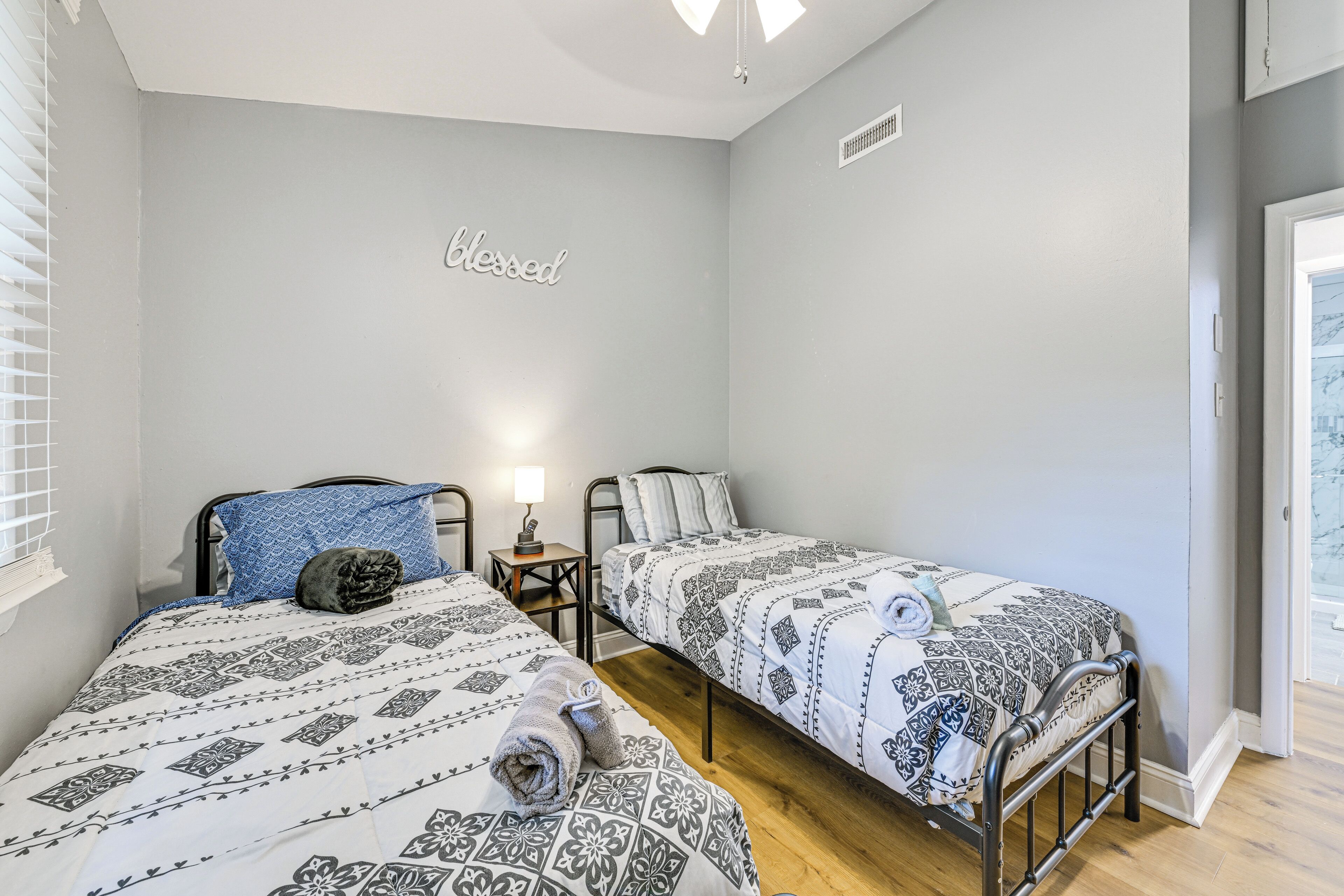 Une chambre avec deux lits simples, une table de chevet avec une lampe et un mur orné d’une inscription « béni ».