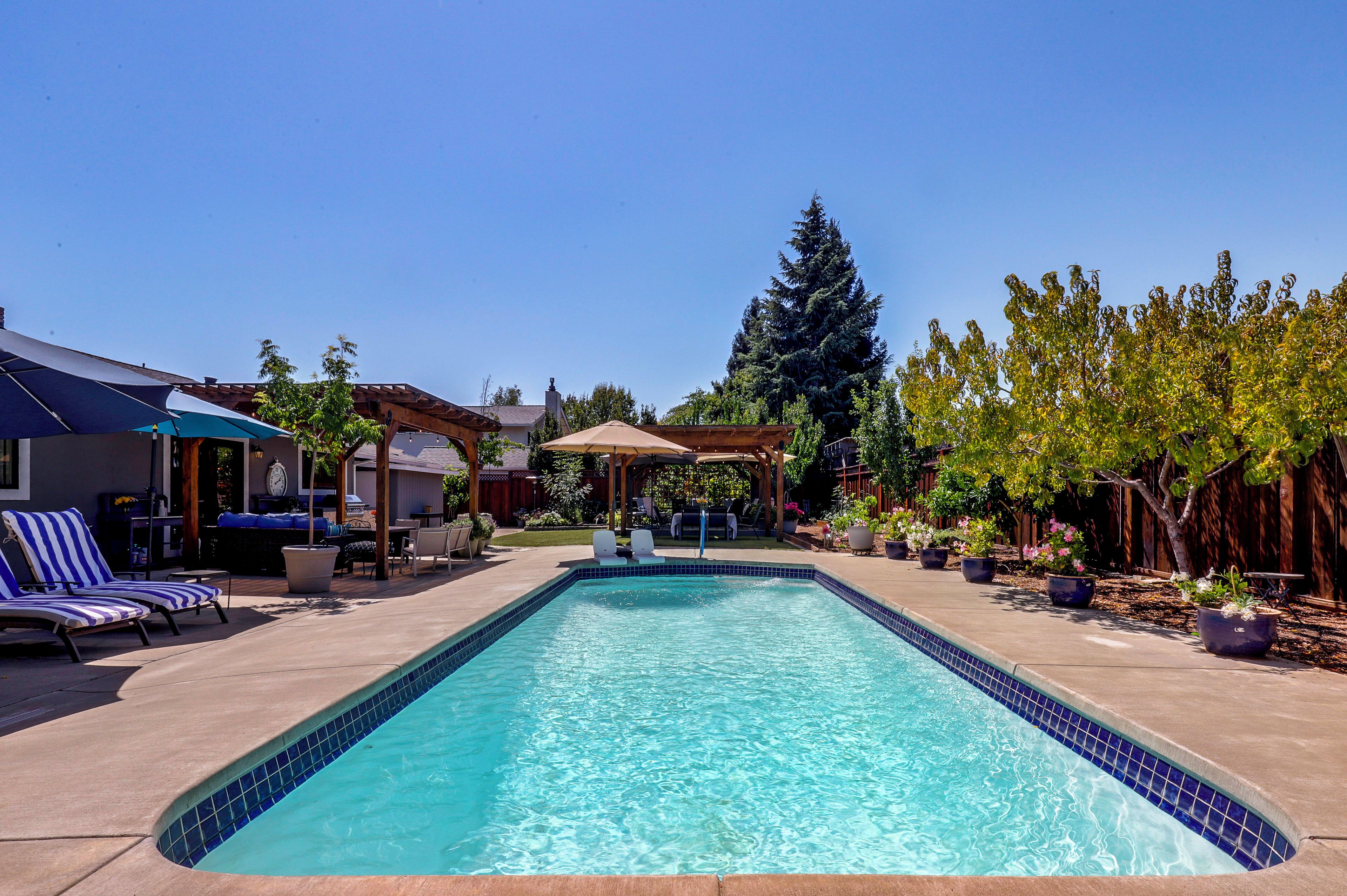 A rectangular swimming pool with a tiled edge, surrounded by lounge chairs and a covered patio area.