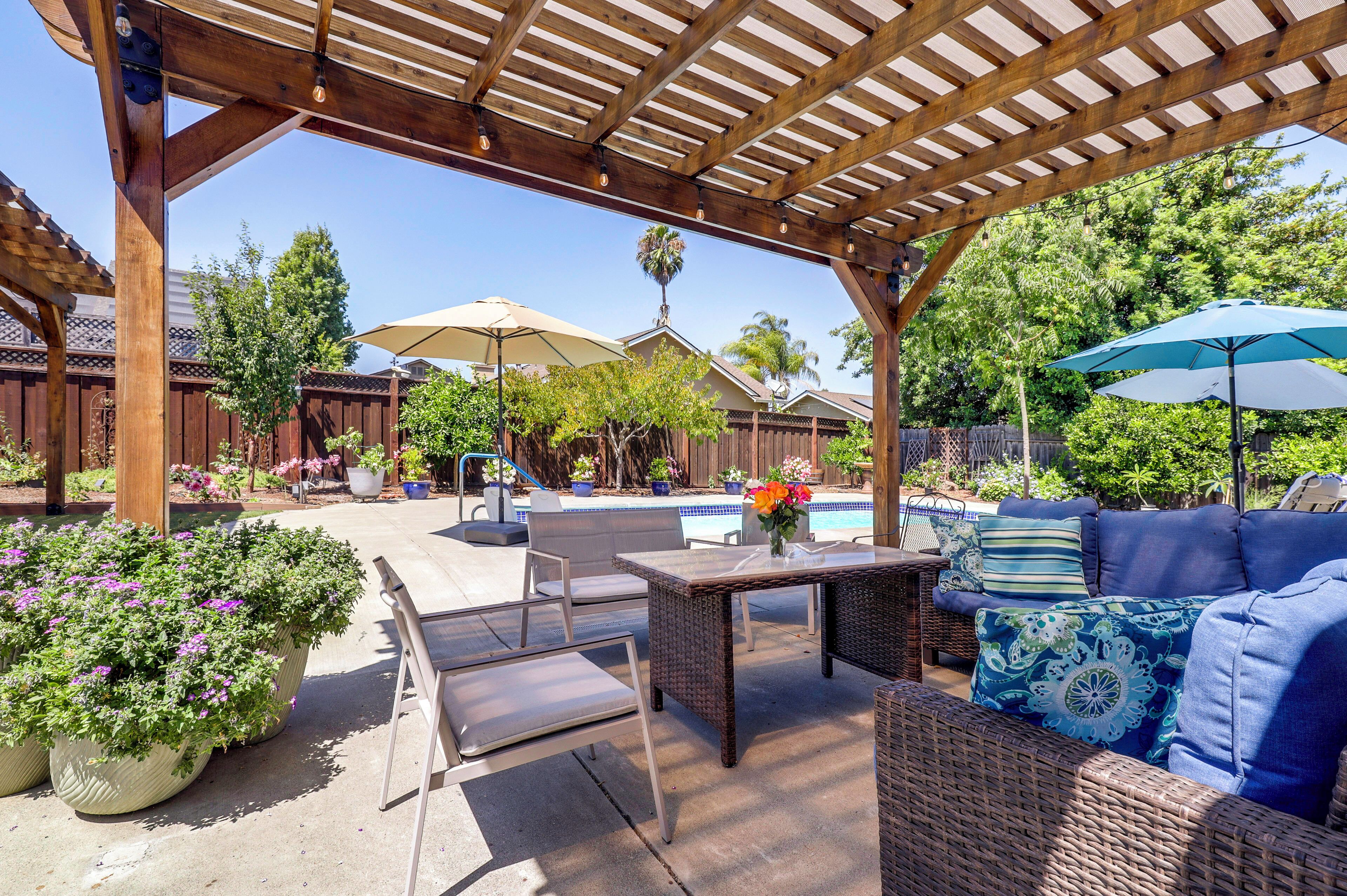 An outdoor patio with a pergola, seating, a pool, and potted plants.
