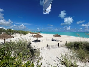 Uma praia com guarda-sóis de palha e espreguiçadeiras, um caminho de areia e um céu azul límpido.