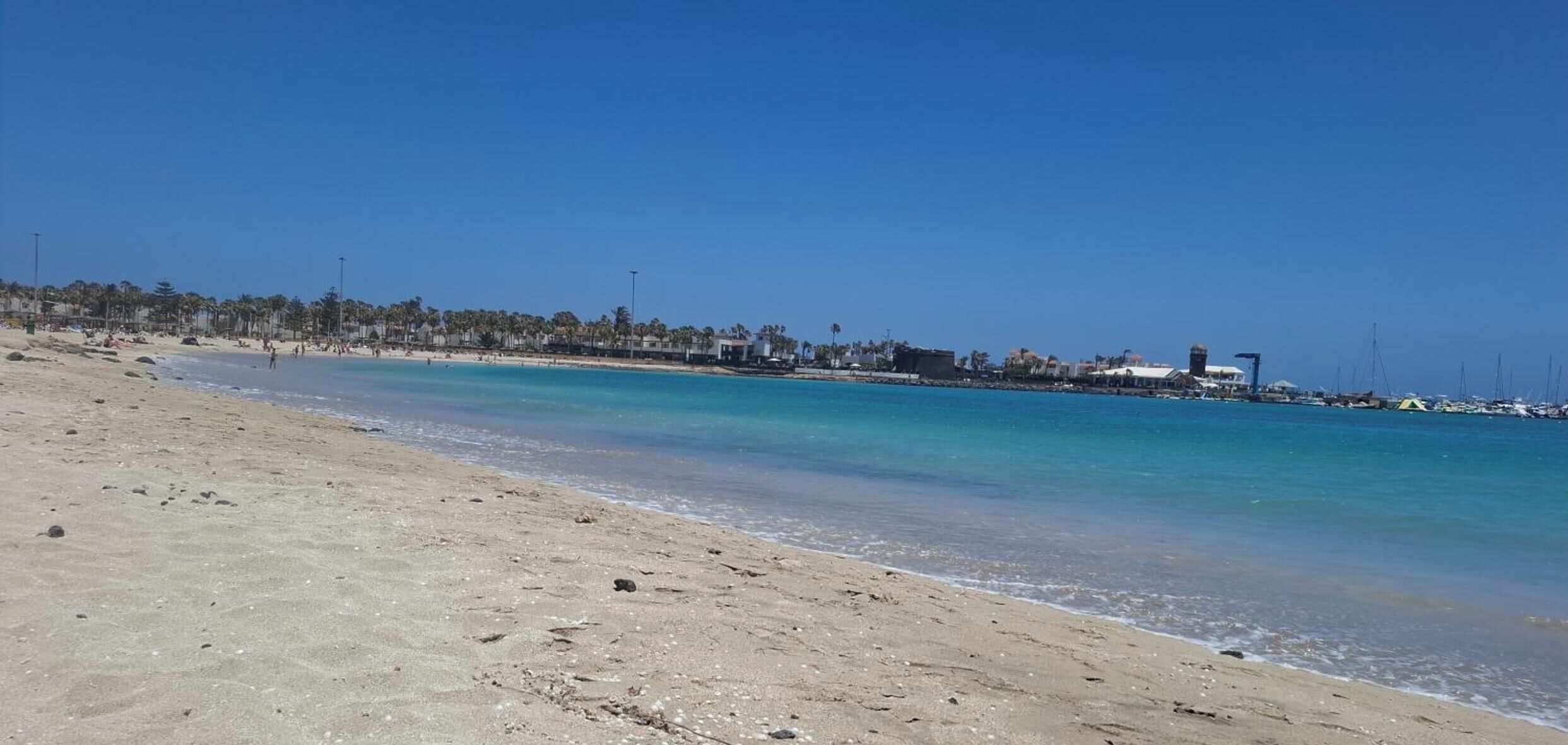 Una playa con agua turquesa cristalina, una orilla arenosa y una hilera de edificios al fondo.