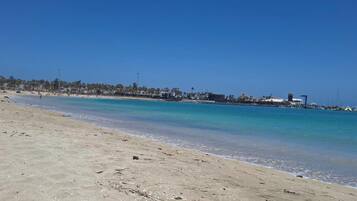 Una playa con agua turquesa cristalina, una orilla arenosa y una hilera de edificios al fondo.
