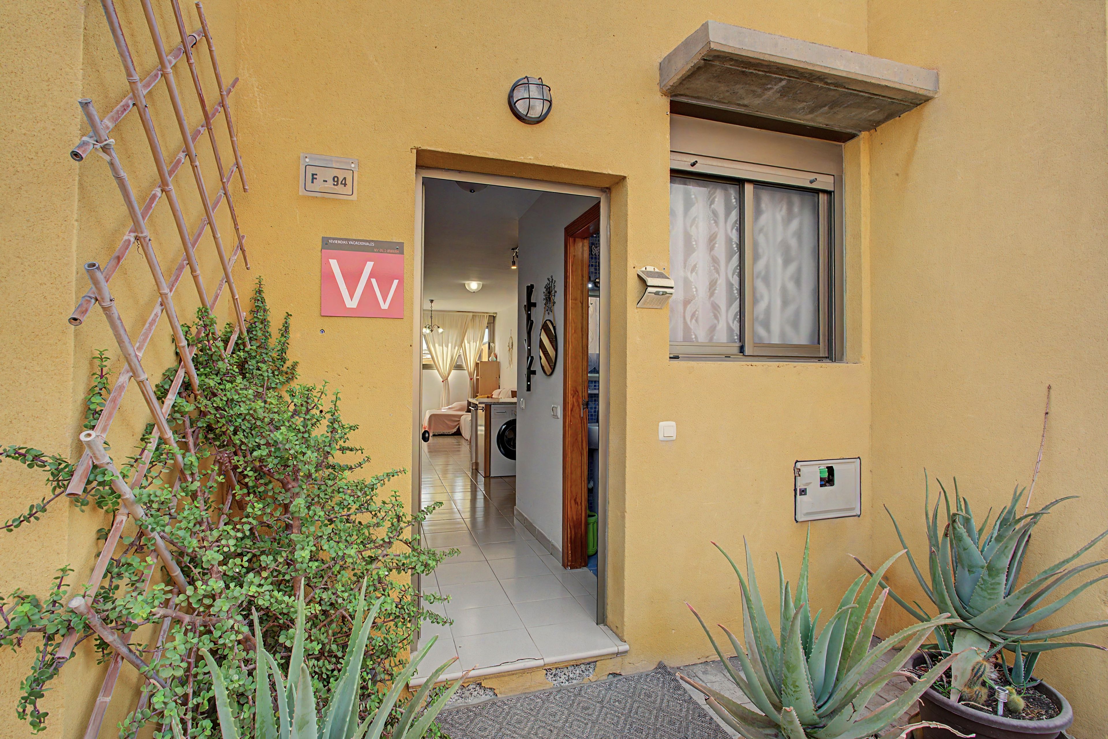 Un edificio amarillo con cerca de bambú, una puerta con un cartel en forma de 'V' y un ventanal con vidrio esmerilado.