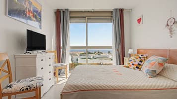 Un dormitorio con cama, cómoda, silla, televisor y ventana con cortinas.