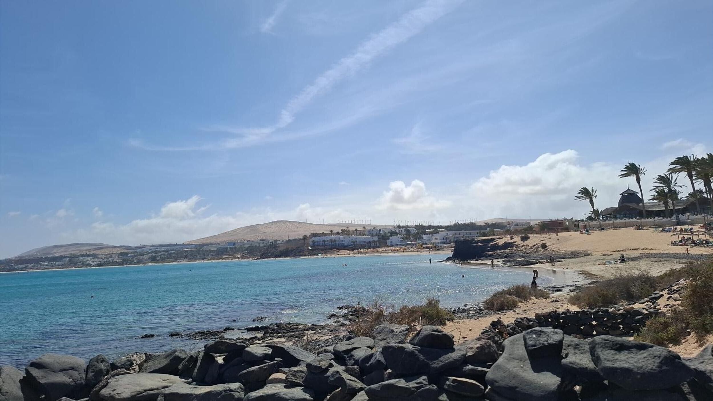 Un paisaje costero con una costa rocosa, mar azul cristalino y una colina distante con edificios.