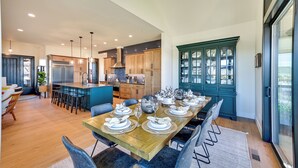 A modern kitchen with a dining area set for a meal, featuring a long wooden table, bar stools, and a built-in island with a sink. - Golf Views, Resort Pool Access! Brasada Ranch Home (Powell Butte)
