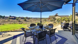 An outdoor patio with a table set for dining, a large umbrella, and a view of a golf course and distant buildings. - Golf Views, Resort Pool Access! Brasada Ranch Home (Powell Butte)