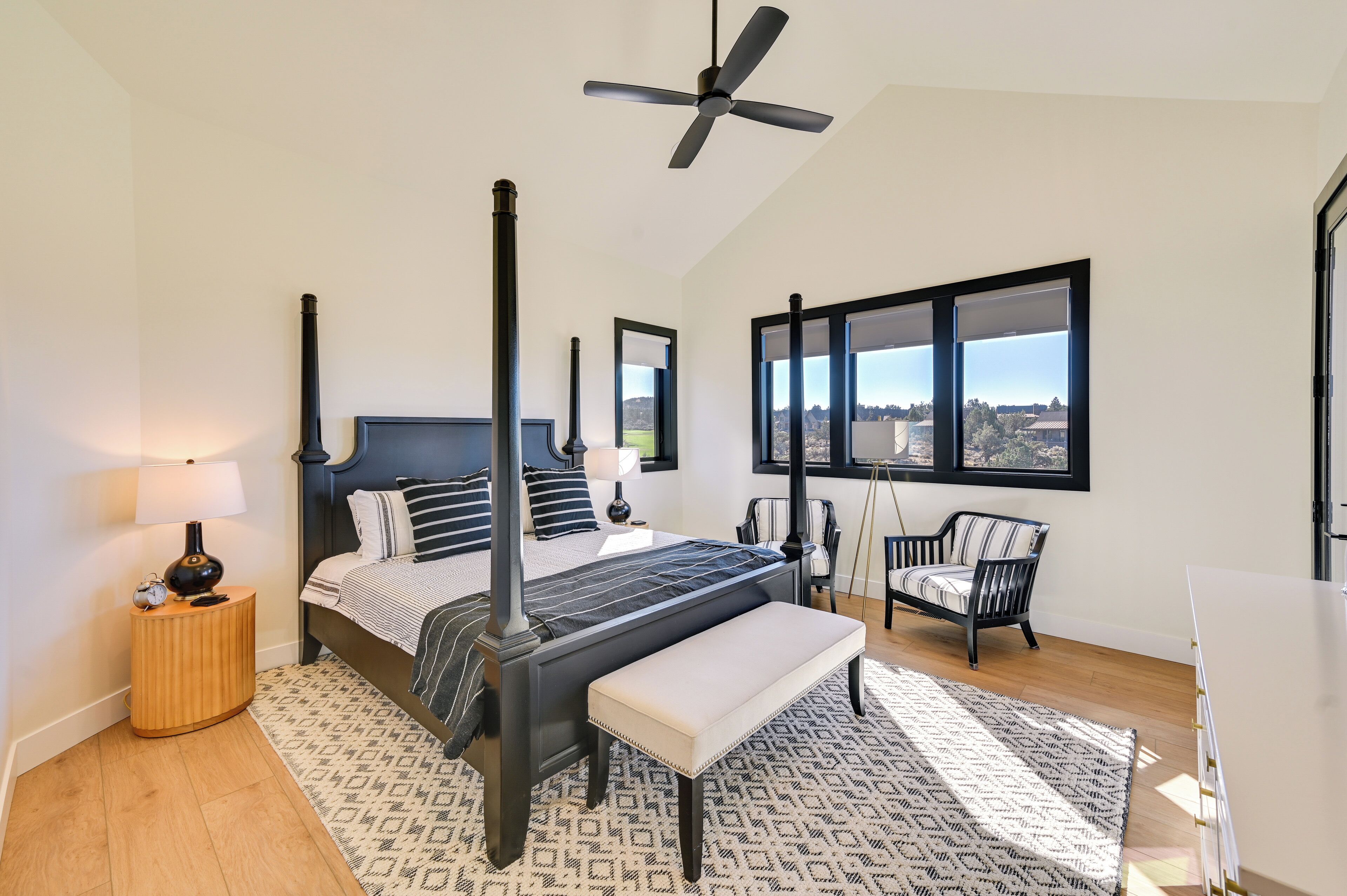 A modern bedroom with a four-poster bed, a standing fan, a large window with a view, a bedside table with a lamp, and a chair.