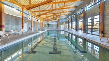 A large indoor swimming pool with a high ceiling and wooden beams, surrounded by seating areas and large windows.