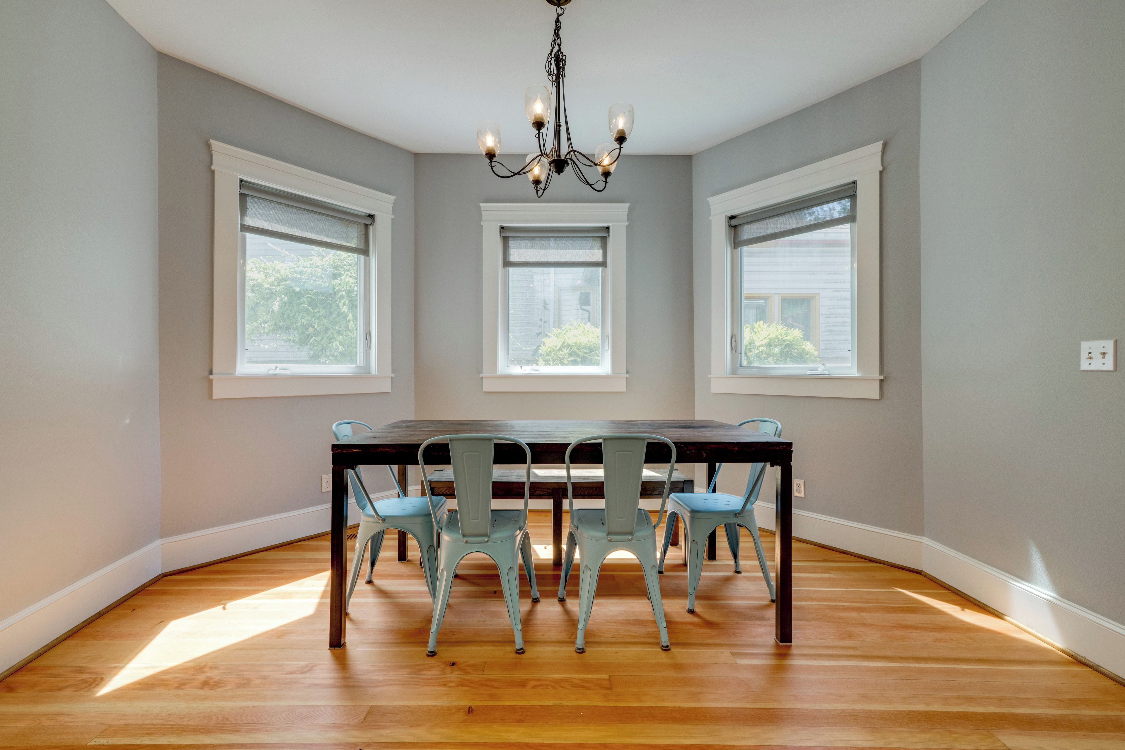 A dining room with a wooden table and four chairs, three windows, and a chandelier.