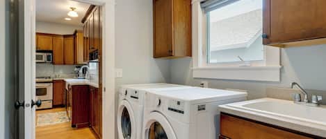 A laundry area with two front-loading washing machines, wooden cabinets, a window with a view, and a kitchen in the background.