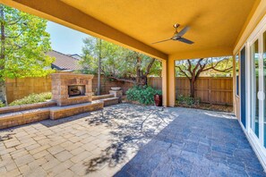 A covered patio with a stone fireplace, a ceiling fan, and a view of a residential area with trees and a fence.