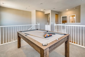 A pool table with balls and cues, surrounded by a white railing, in a room with a neutral color scheme and recessed lighting.