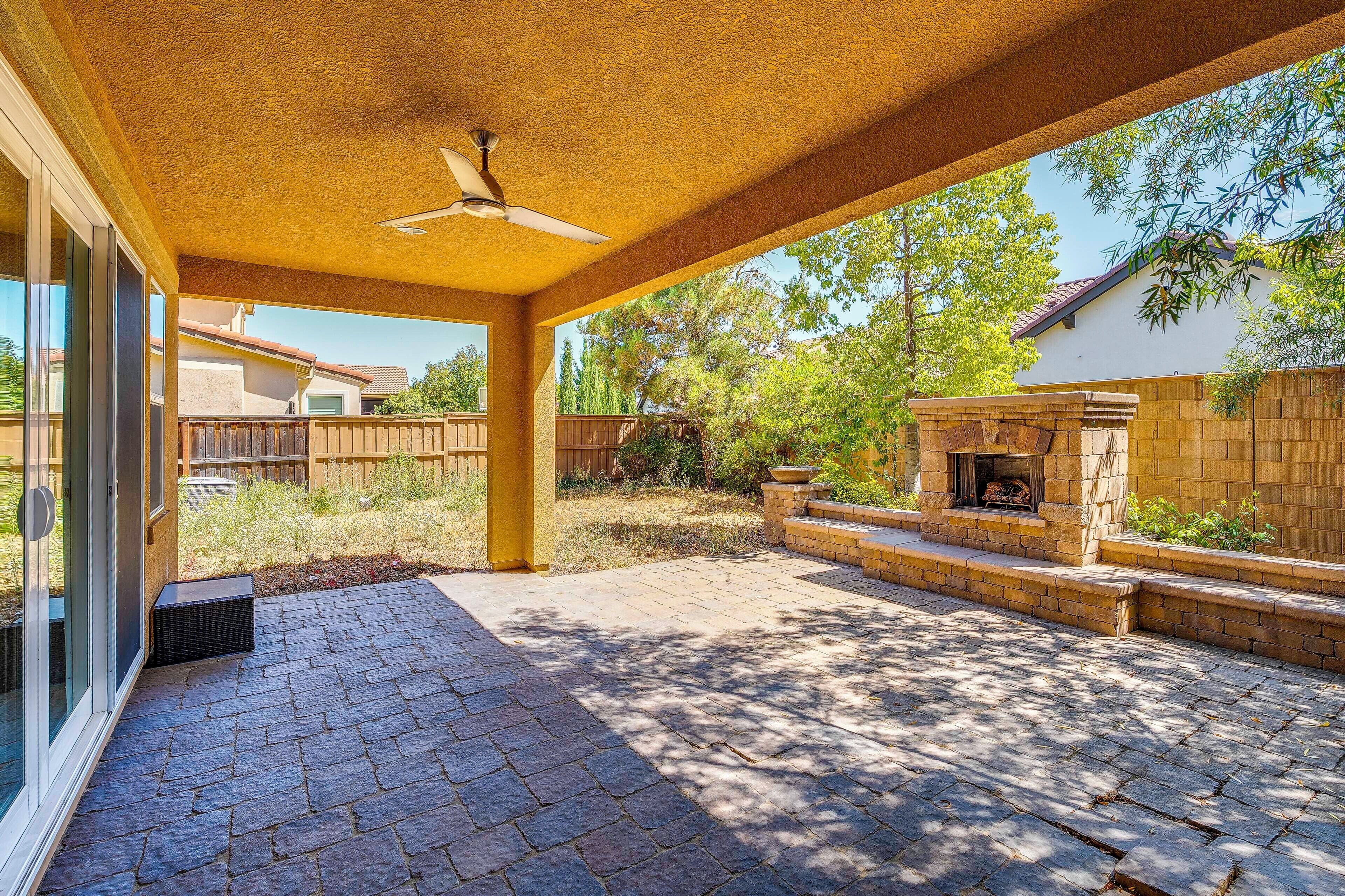 A covered patio area with a stone fireplace, a ceiling fan, and a view of a residential neighborhood.