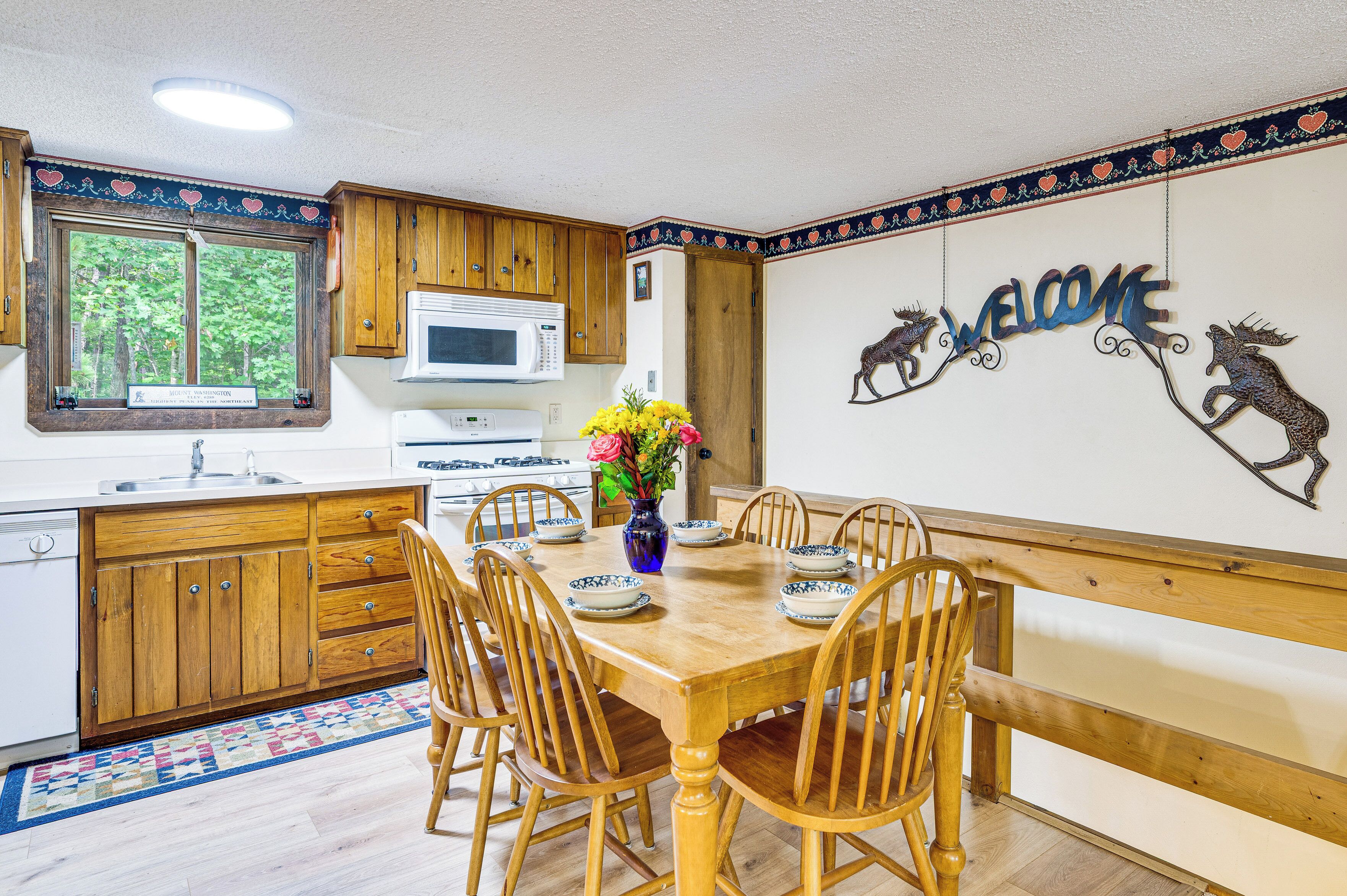 A kitchen with wooden cabinets, a microwave, and a stove. There is a dining table with chairs and a vase of flowers. A welcome sign with animal silhouettes is on the wall.