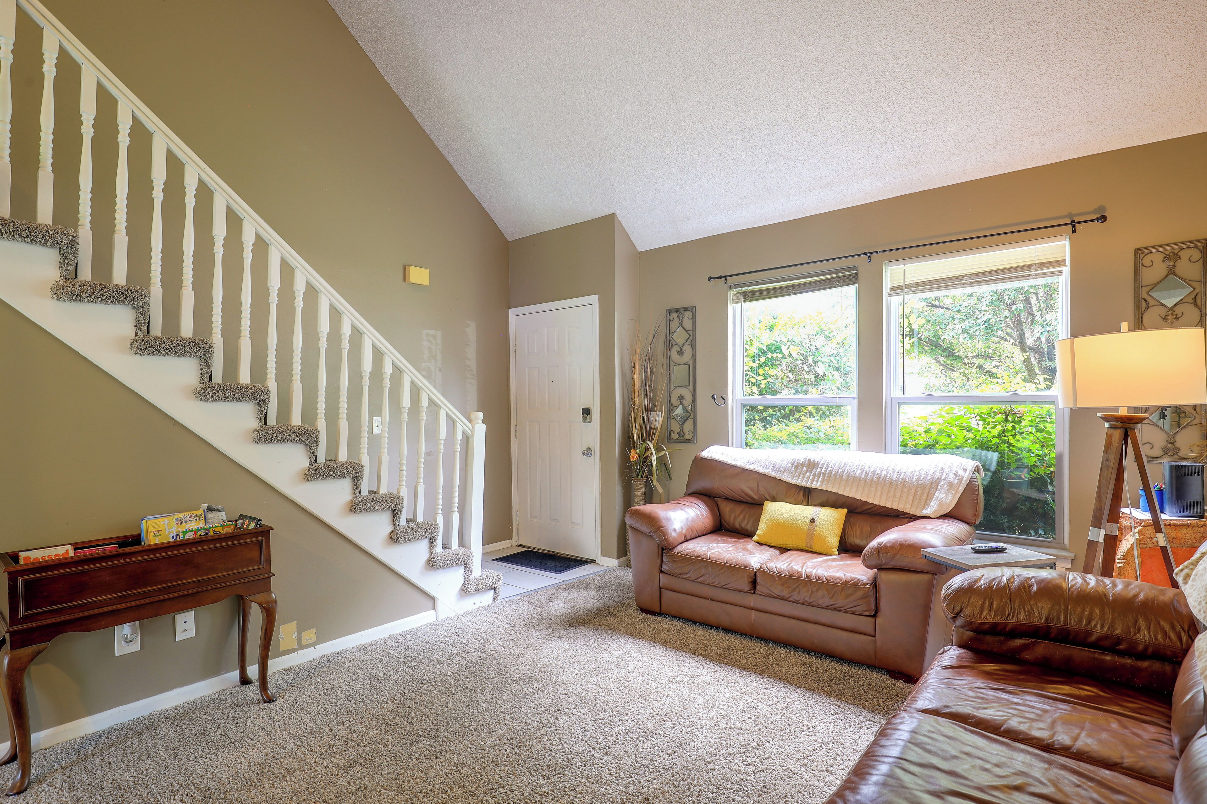 A living room with a staircase, a brown leather sofa, a coffee table, and a window with a view of greenery.