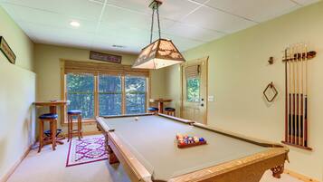 A pool table with a cue rack, two stools, and a window with blinds.
