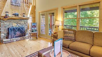 A cozy cabin interior with a stone fireplace, a wooden staircase, a comfortable sofa, and large windows offering a view of the outdoors.