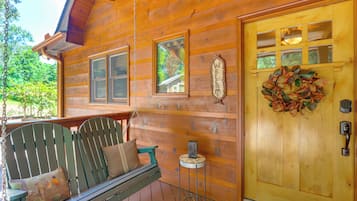 A wooden cabin with a porch, a swing chair, a door with a wreath, and a small table with a decorative item.