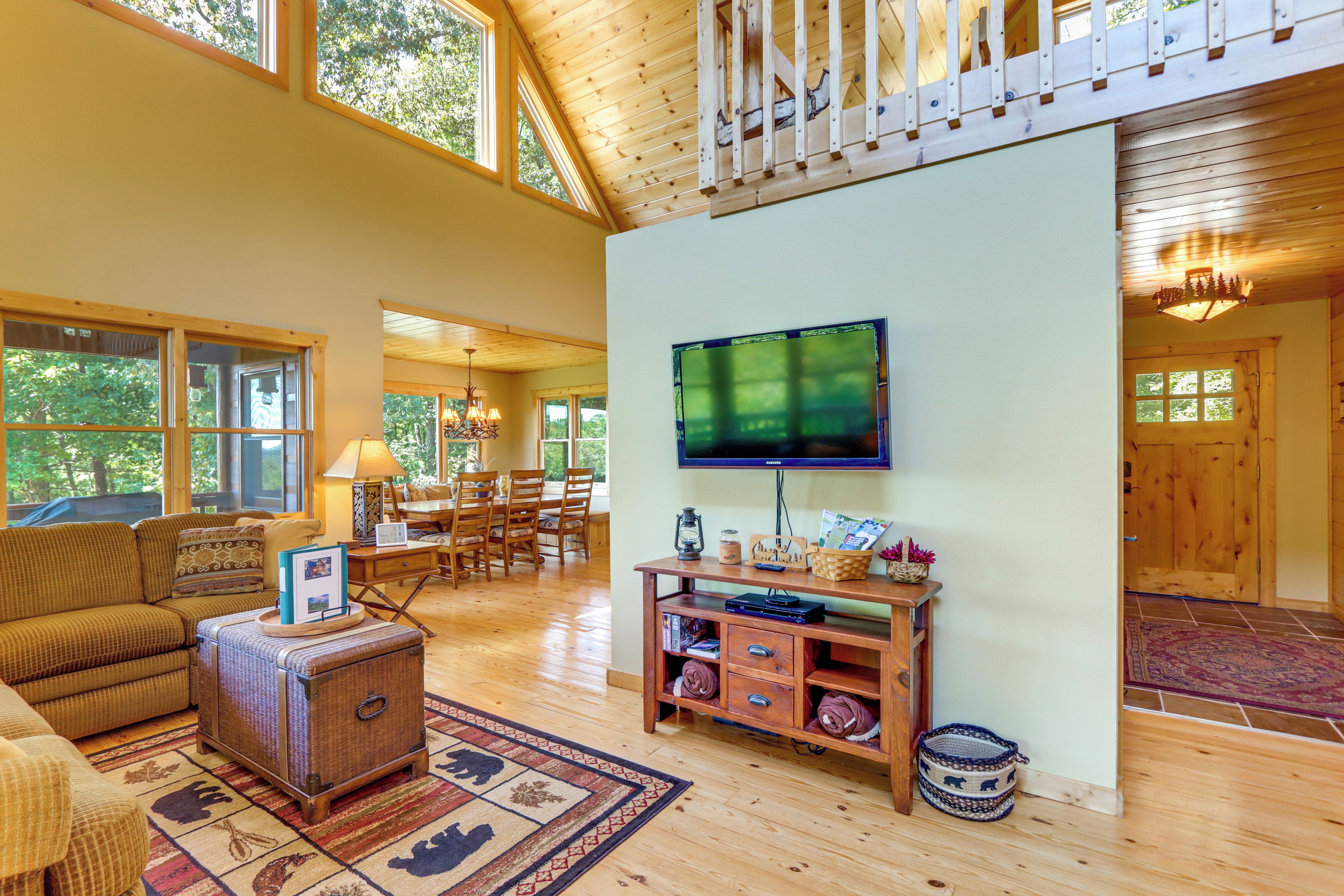 A spacious living room with a large television mounted on the wall, wooden flooring, and a high ceiling with exposed beams.