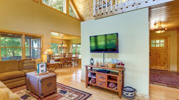 A spacious living room with a large television mounted on the wall, wooden flooring, and a high ceiling with exposed beams.