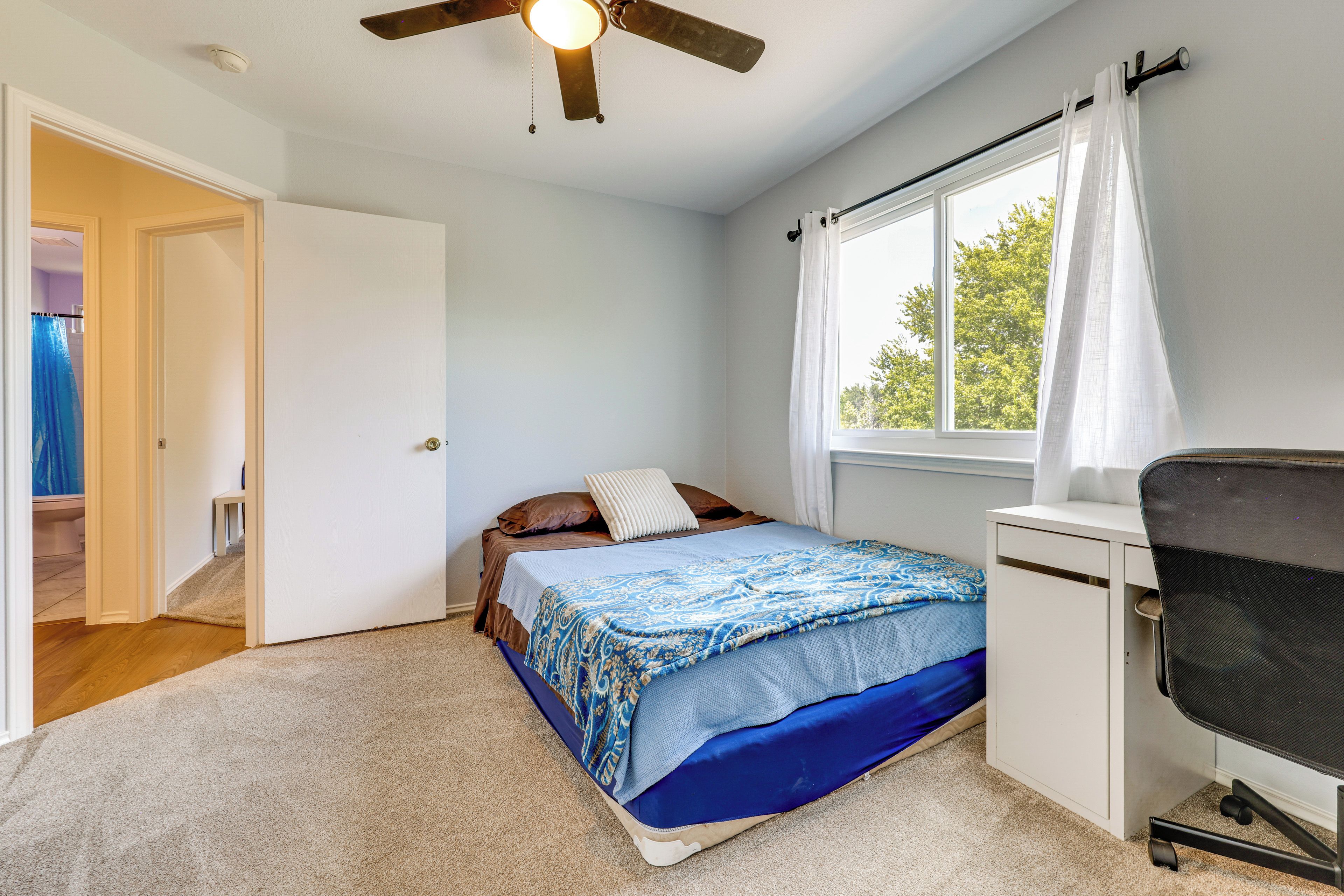 A bedroom with a bed, a desk, a chair, a window with curtains, and a ceiling fan.