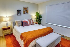 Un dormitorio con cama, mesitas de noche, lámpara, ventana con persianas y una planta.