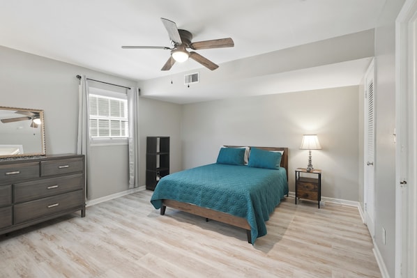 A bedroom with a bed, a dresser, a ceiling fan, a window with curtains, and a nightstand with a lamp.