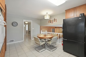 A kitchen with a black refrigerator, a dining table with chairs, a clock on the wall, and wooden cabinets.