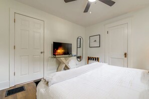 Une chambre à coucher avec un lit, un téléviseur posé sur une table en verre, un ventilateur de plafond et des murs blancs.