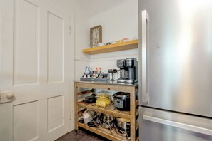 A compact kitchen with a stainless steel refrigerator, a coffee maker, and a shelf with various kitchen utensils.